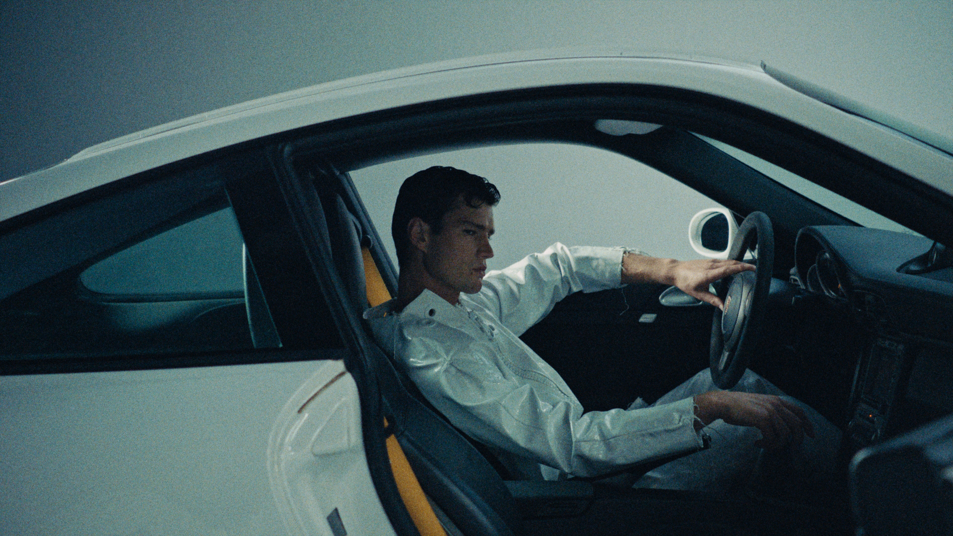 Model in a white leather outfit with his hand on the wheel.